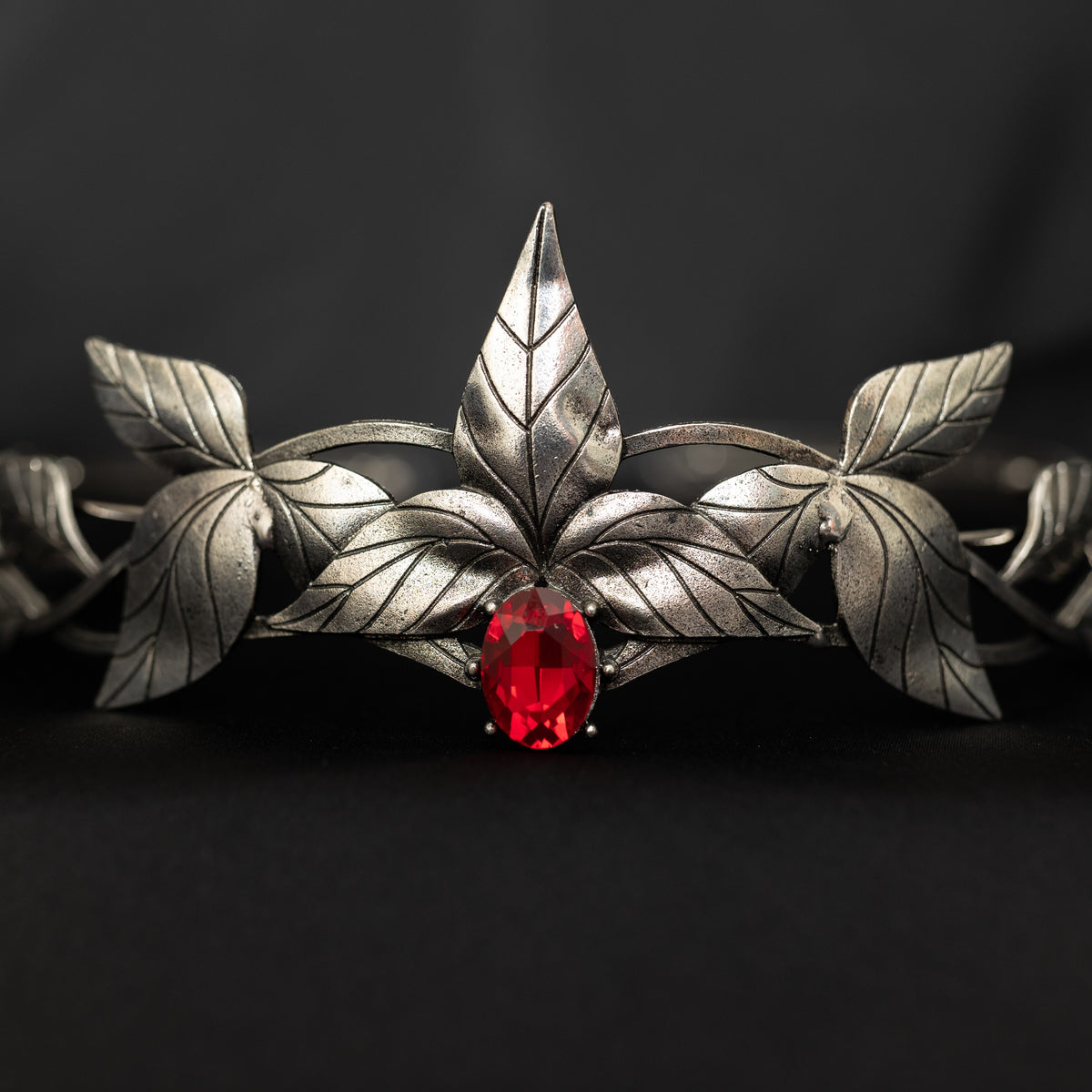 A silver-colored tiara with laurel leaf patterns and a central red stone, presented against a black background. Front view 