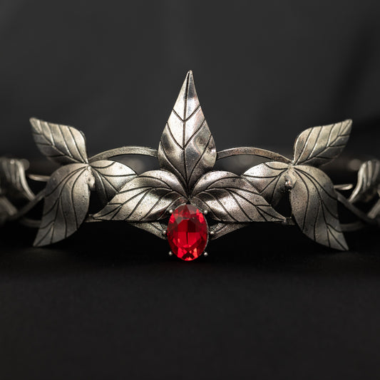 A silver-colored tiara with laurel leaf patterns and a central red stone, presented against a black background. Front view 