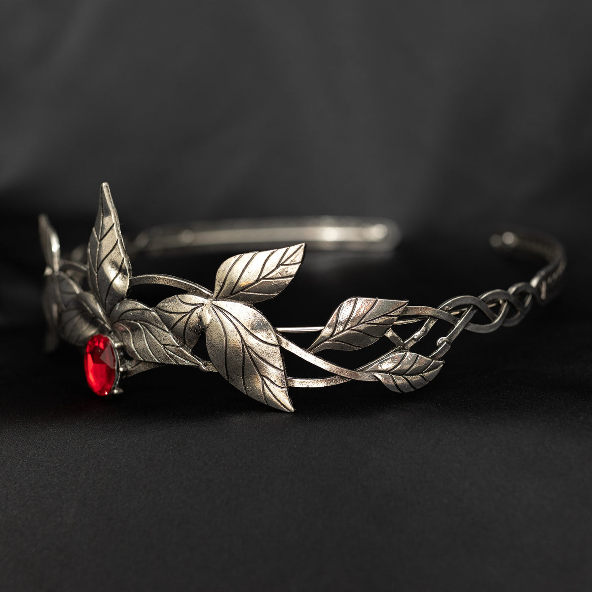 A silver-colored tiara with laurel leaf patterns and a central red stone, presented against a black background. Front side view 