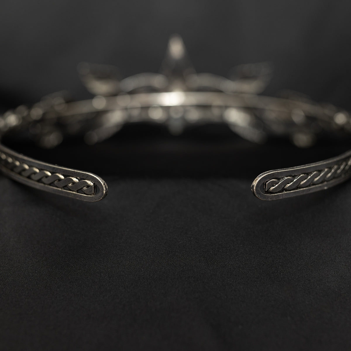 A silver-colored tiara with laurel leaf patterns and a central red stone, presented against a black background. Rear view 