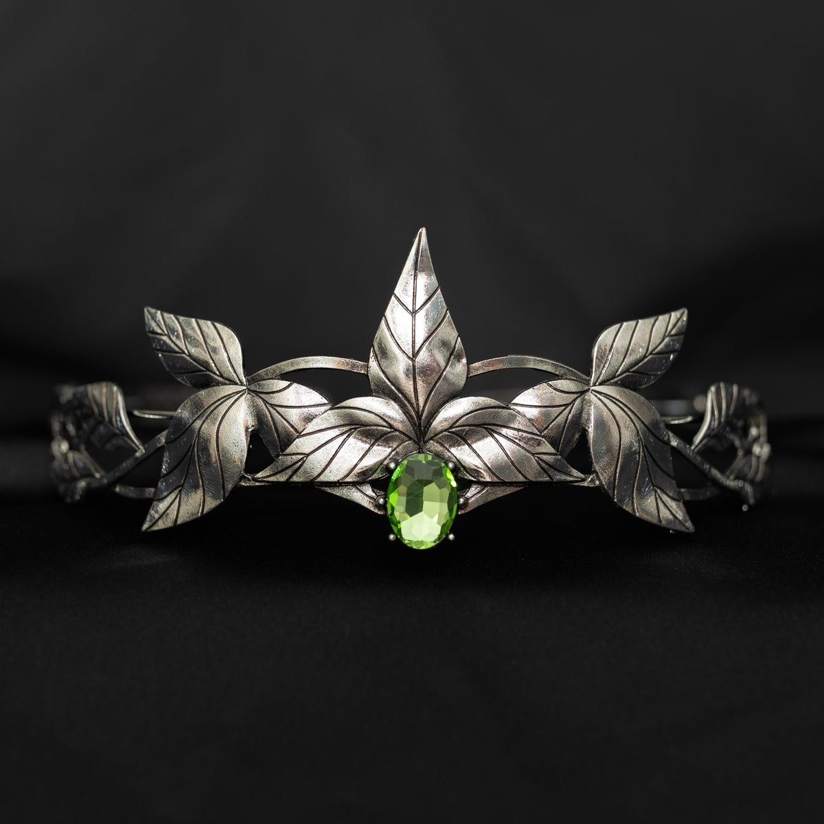 A silver-colored tiara with laurel leaf patterns and a central light green gemstone, displayed against a black background. Front view 