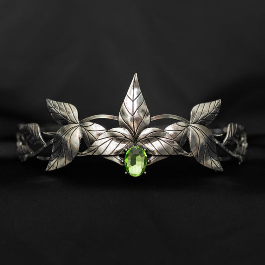 A silver-colored tiara with laurel leaf patterns and a central light green gemstone, displayed against a black background. Front view 