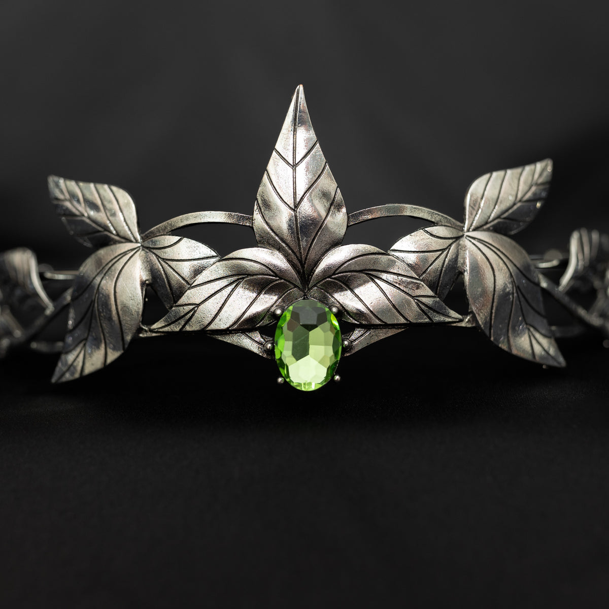 A silver-colored tiara with laurel leaf patterns and a central light green gemstone, displayed against a black background. Front close up view 