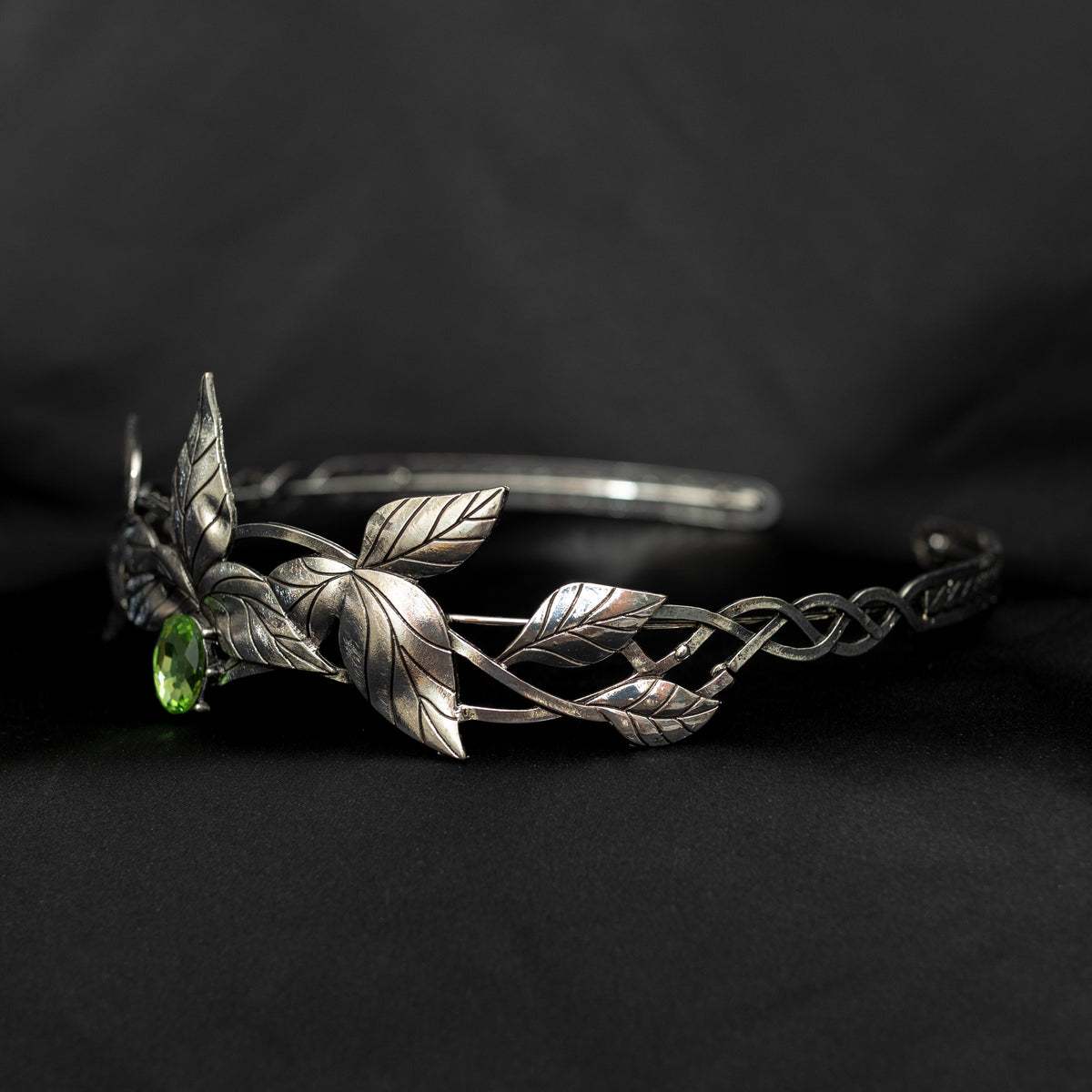 A silver-colored tiara with laurel leaf patterns and a central light green gemstone, displayed against a black background. Side front view 