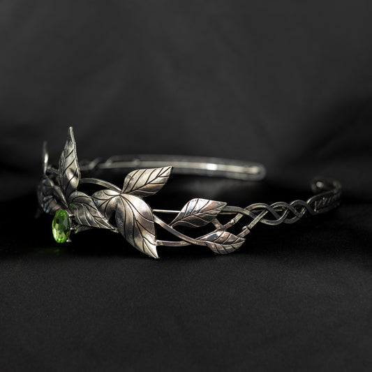 A silver-colored tiara with laurel leaf patterns and a central light green gemstone, displayed against a black background. Side front view 