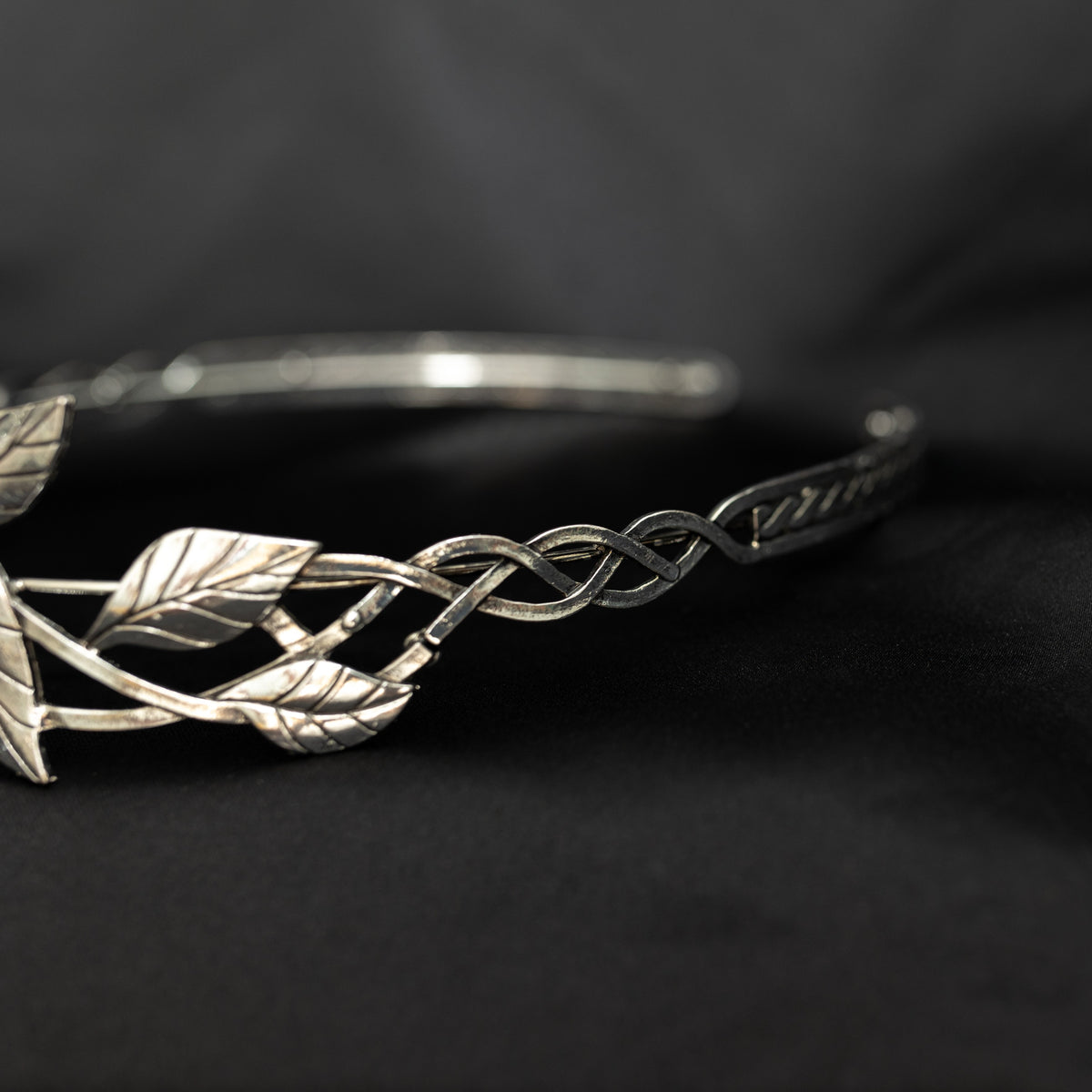 A silver-colored tiara with laurel leaf patterns and a central light green gemstone, displayed against a black background. Side view 