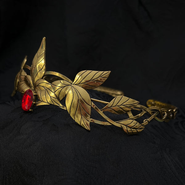 A vintage gold-toned brooch featuring a leafy vine design and a small red oval stone, displayed on a textured black fabric background.
