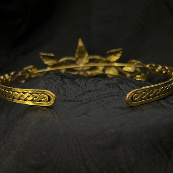 An ornate gold-toned metal headband with decorative scrollwork and a leaf-like crest at the top, displayed on a dark textured fabric background.