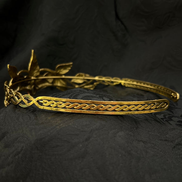 A gold-toned metal tiara with leaf and vine motifs, displayed on a textured black fabric background.