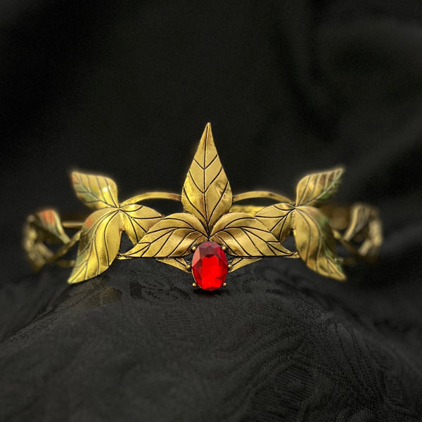 A gold-toned leaf-shaped cuff bracelet with a central red gemstone, displayed on a textured black fabric background.