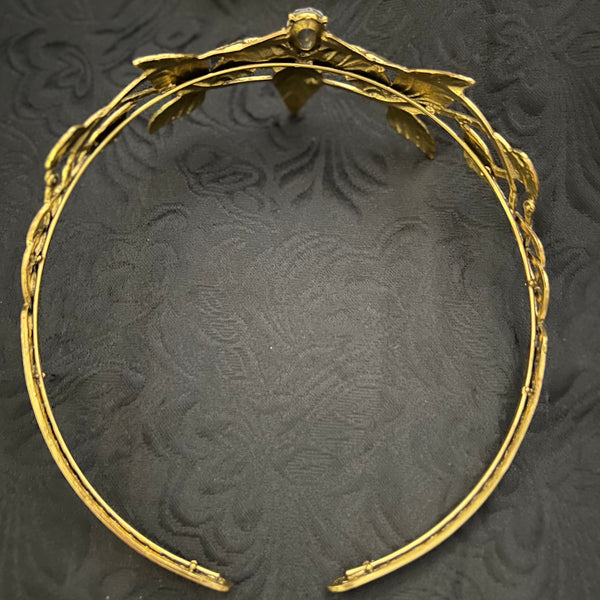 A gold-toned metal headband with a decorative leafy crown design and a small central embellishment, photographed on a dark textured fabric background.