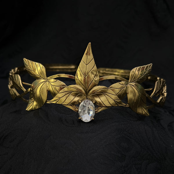 A vintage gold-toned brooch featuring sculpted leaves and a central clear oval gemstone, displayed on a textured black fabric background.
