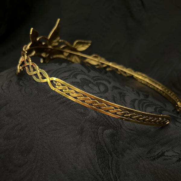A gold-toned metal crown with ornate leaf and knot motifs, displayed on a dark textured fabric background.