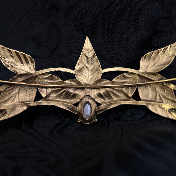 A close-up of a vintage gold-toned brooch featuring leaf motifs and a central oval blue-gray stone, set against a textured black fabric background.