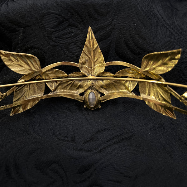 A gold-toned metal brooch shaped like a leafy vine with a small oval dark stone, displayed on a textured black fabric background.
