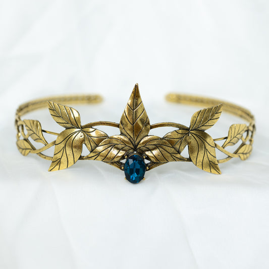 A golden tiara crown featuring a central dark blue stone, surrounded by laurel leaf detailing on a white background. Front view 