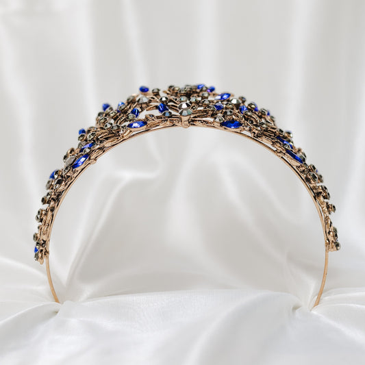 A tiara featuring a baroque-inspired design with blue and gray crystals, antique gold-colored metal, and loops on each end for securing to hair._Bottom
