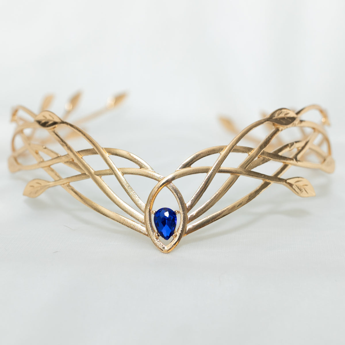 A gold-colored metal crown with a woven design and a central blue crystal, displayed against a white background.