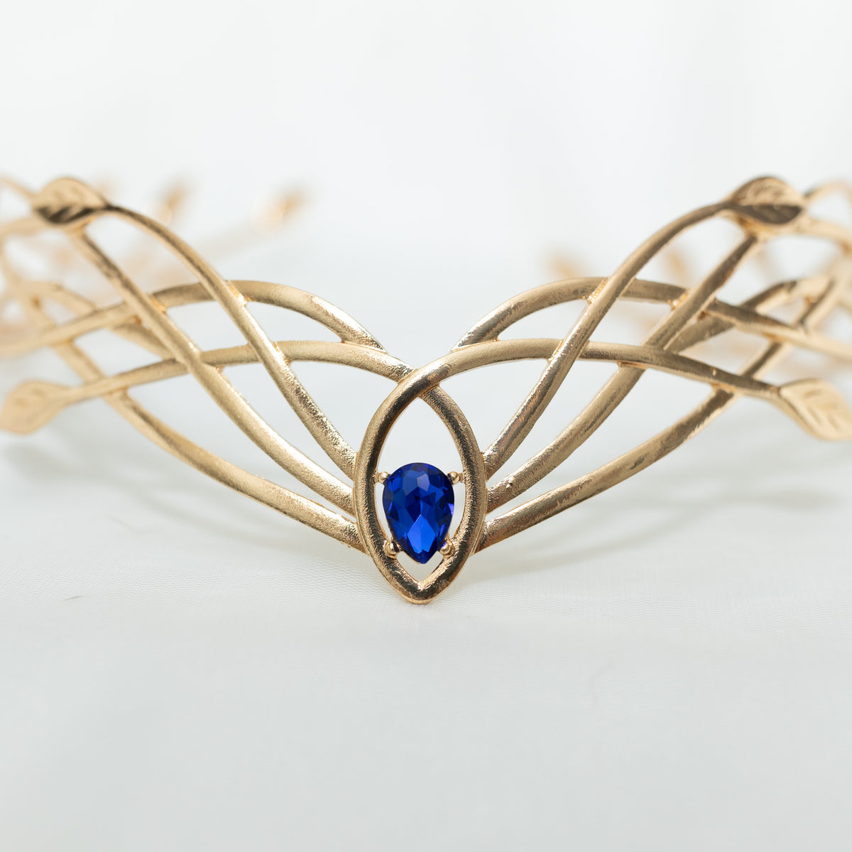 A gold-colored metal crown with a woven design and a central blue crystal, displayed against a white background.