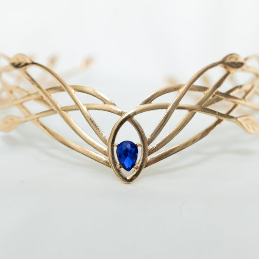 A gold-colored metal crown with a woven design and a central blue crystal, displayed against a white background.