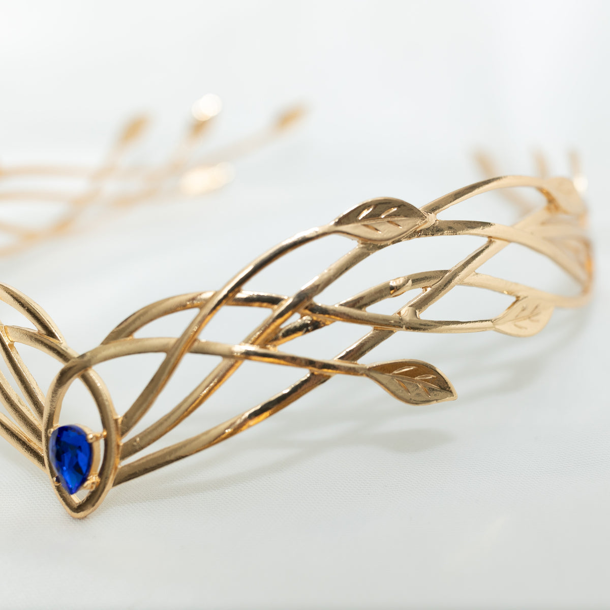 A gold-colored metal crown with a woven design and a central blue crystal, displayed against a white background.