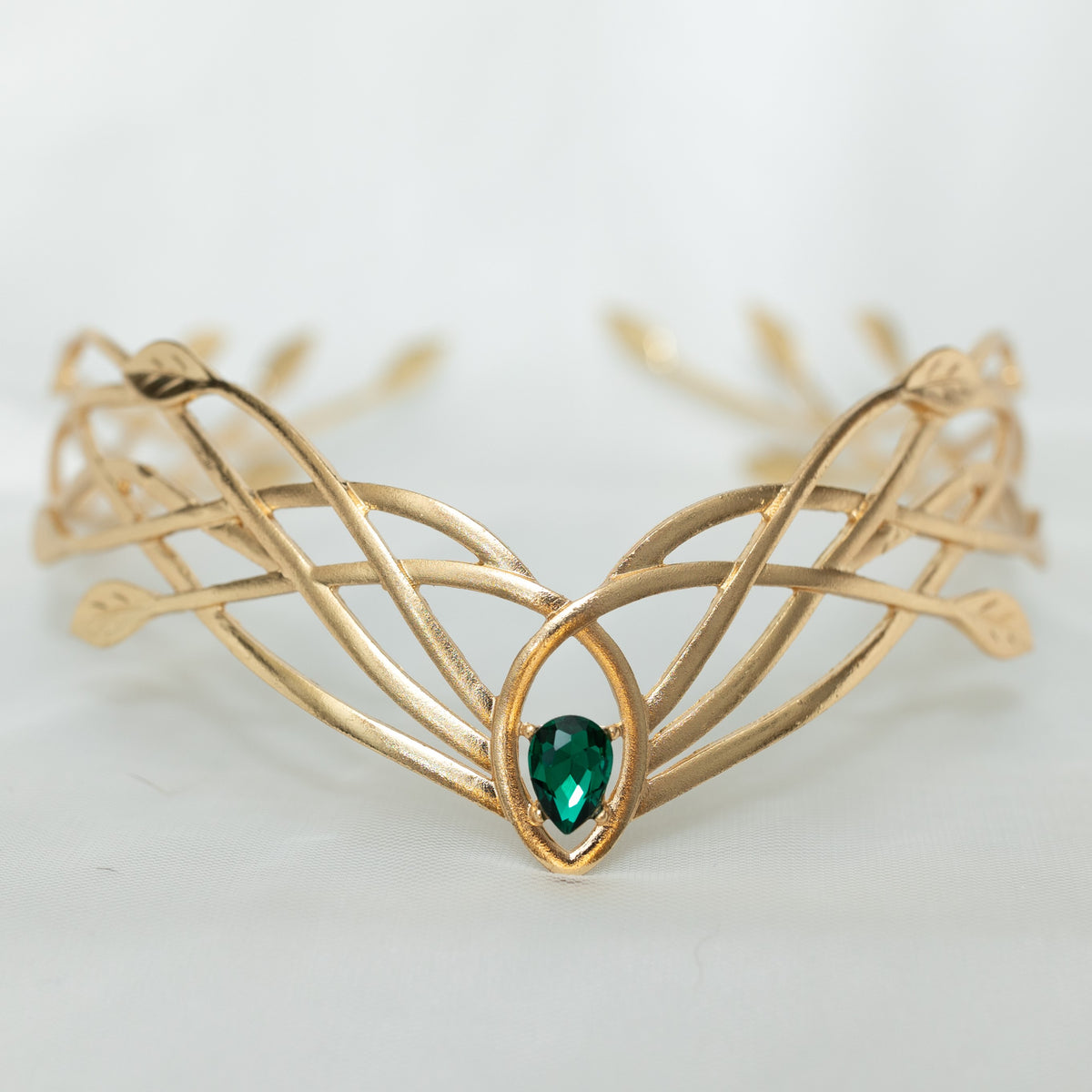 A gold-colored metal crown with a woven design and a central green crystal, displayed against a white background.