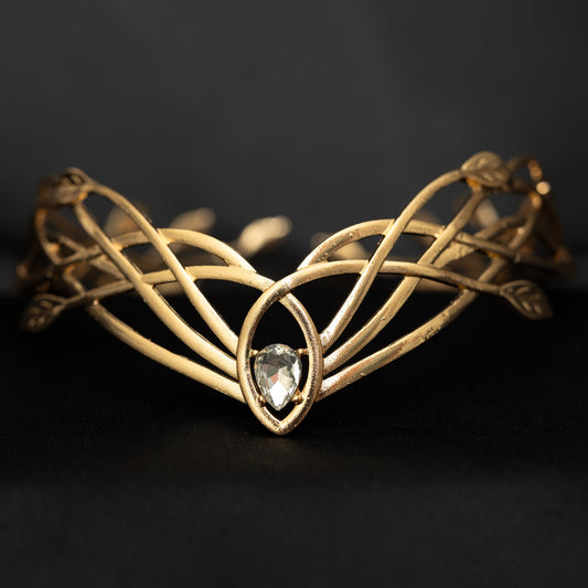 A gold-colored metal crown with a woven design and a central clear crystal, displayed against a black background.