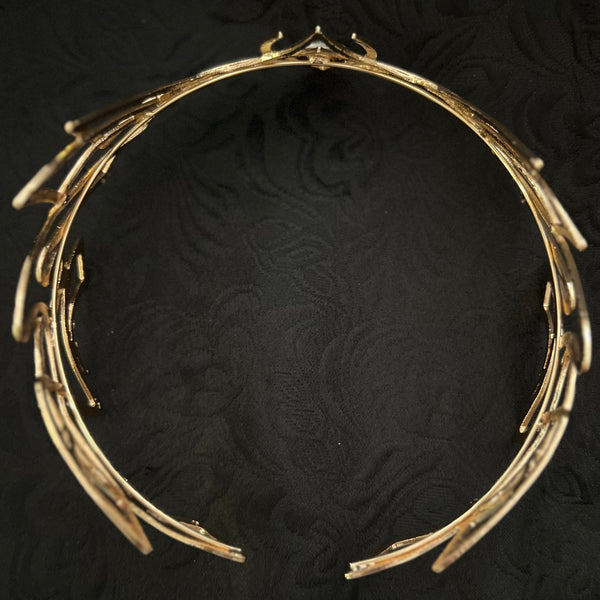 A gold-toned metallic headband with a branch-like organic design, displayed on a textured black fabric background.