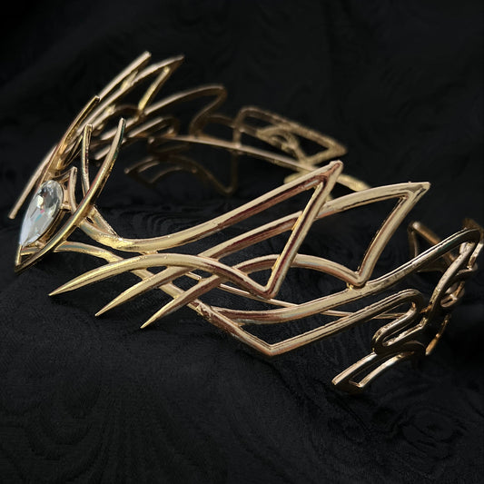 A gold-toned geometric metal brooch with sharp angular lines and a clear faceted gemstone, displayed on a textured black fabric background.