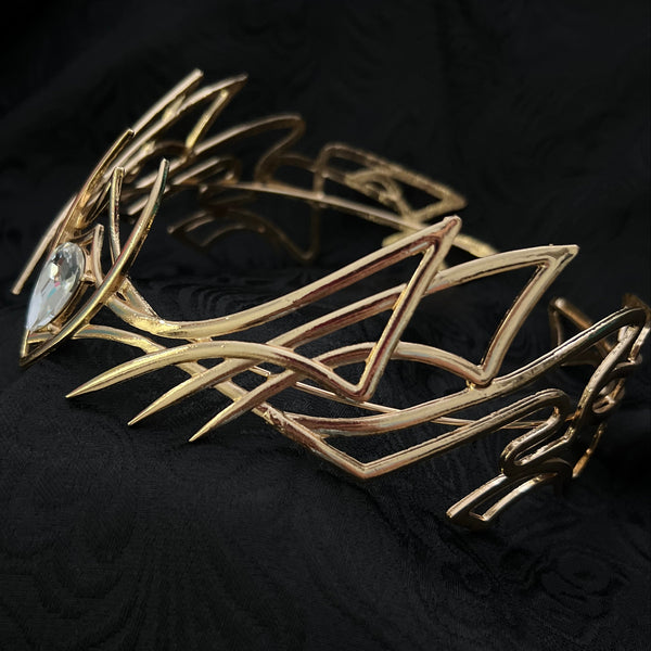 A gold-toned geometric metal brooch with sharp angular lines and a clear faceted gemstone, displayed on a textured black fabric background.