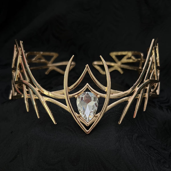 A gold-toned cuff bracelet featuring an intricate, branching organic design and a central clear teardrop gemstone, displayed on a textured black fabric background.