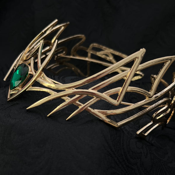 A close-up of a gold-toned abstract geometric brooch with a green faceted gemstone, set against a textured black fabric background.