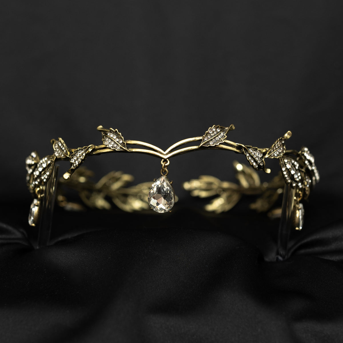 An antique gold circlet crown with leaf details and a crystal drop at the center, displayed on a black background.