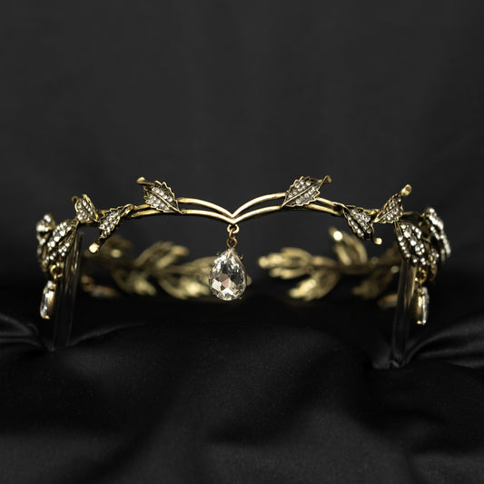 An antique gold circlet crown with leaf details and a crystal drop at the center, displayed on a black background.