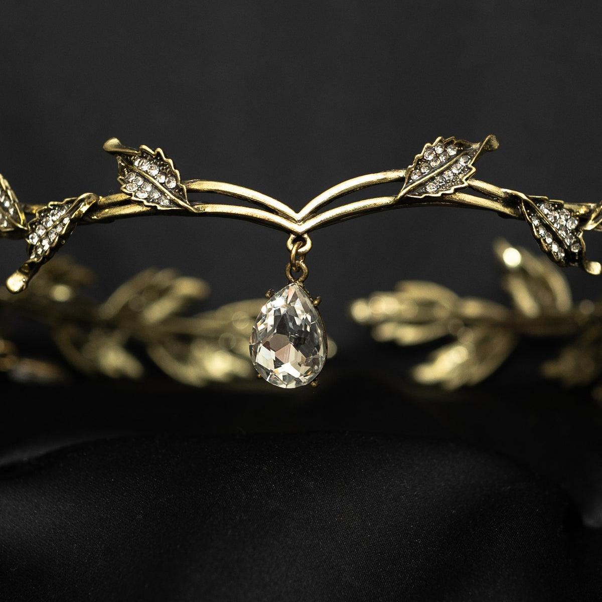 An antique gold circlet crown with leaf details and a crystal drop at the center, displayed on a black background.