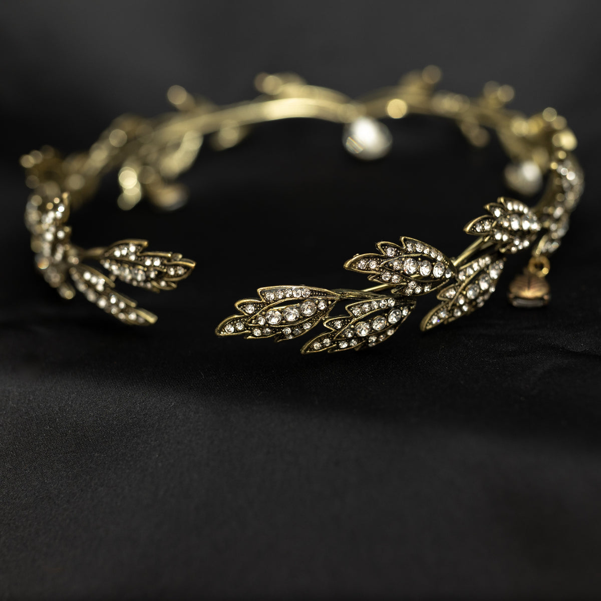 An antique gold circlet crown with leaf details and a crystal drop at the center, displayed on a black background.