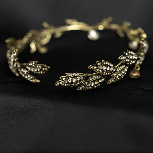 An antique gold circlet crown with leaf details and a crystal drop at the center, displayed on a black background.