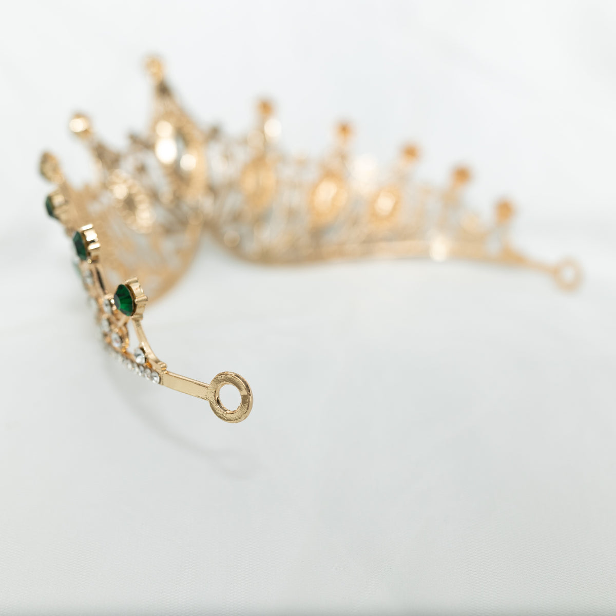 A golden tiara adorned with large green marquise shaped crystals and small clear round crystals, set against a white background. End view 