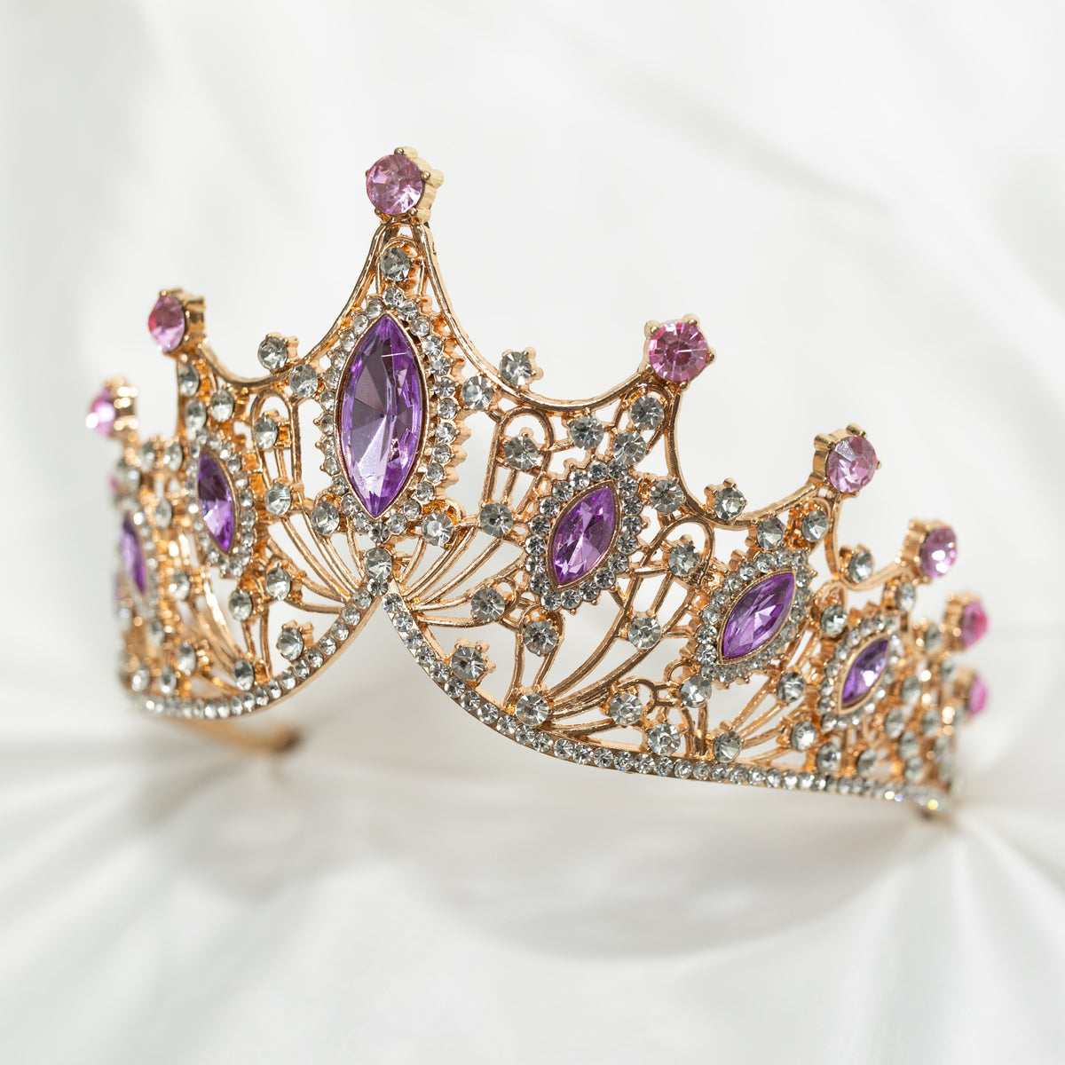 A Lavender and Gold tiara with marquise shaped crystals and clear round crystal detailing on a gold-colored metal structure, displayed against a white fabric background. Front side view 