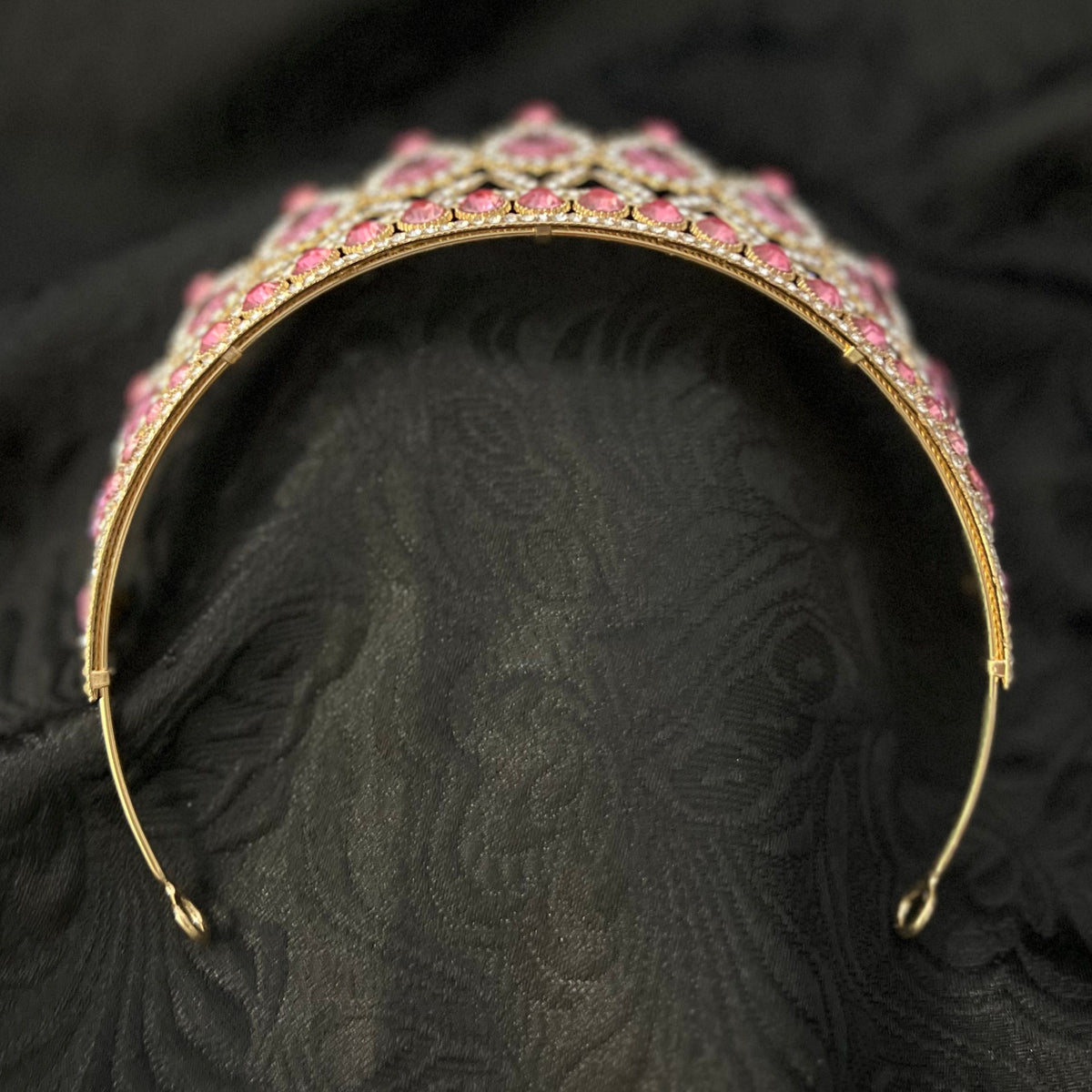 A gold-plated metal tiara headband embellished with pink rhinestones, displayed on a textured black fabric background.
