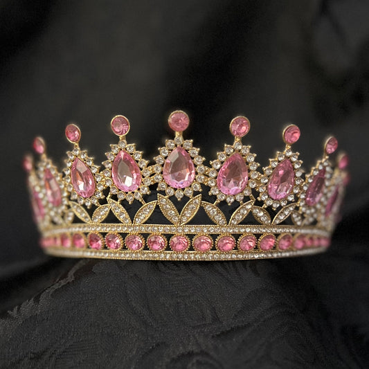 A gold-toned tiara crown adorned with pink gemstones, resting on a draped black fabric background.