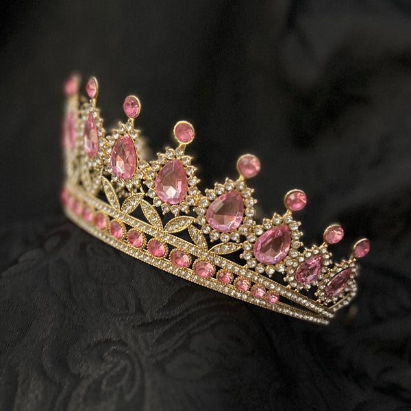 A gold tiara crown embellished with pink gemstones, resting on a textured black fabric background.