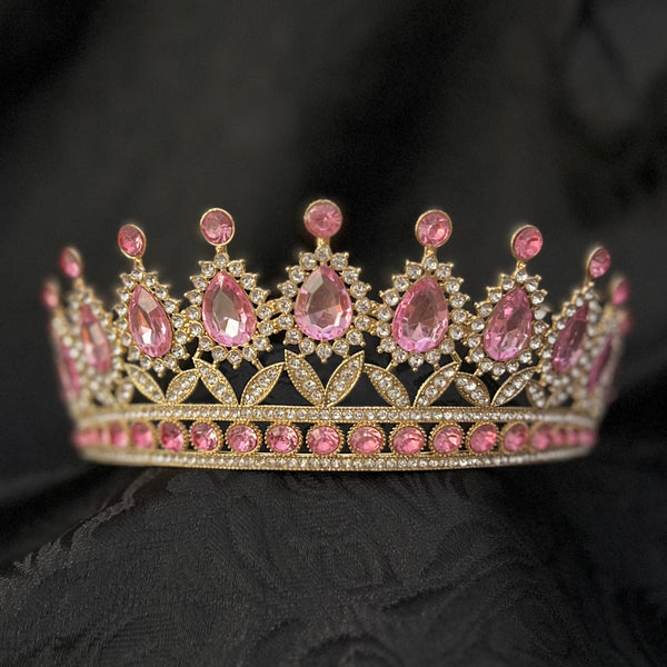 A gold-toned tiara crown adorned with pink gemstones, resting on a draped black fabric background.