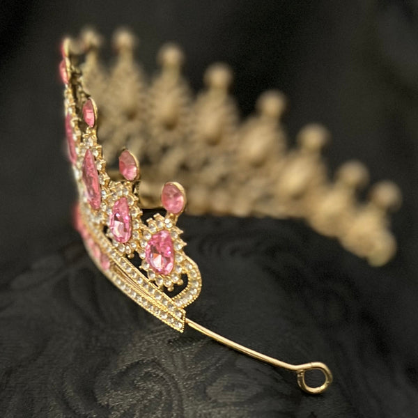 A gold tiara with pink gemstones resting on a textured black fabric background.