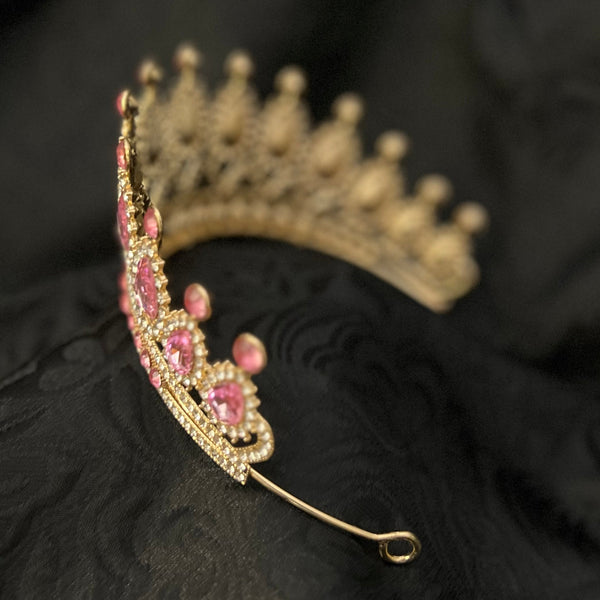 A gold tiara with pink gemstones and clear rhinestones, resting on a textured black fabric background.