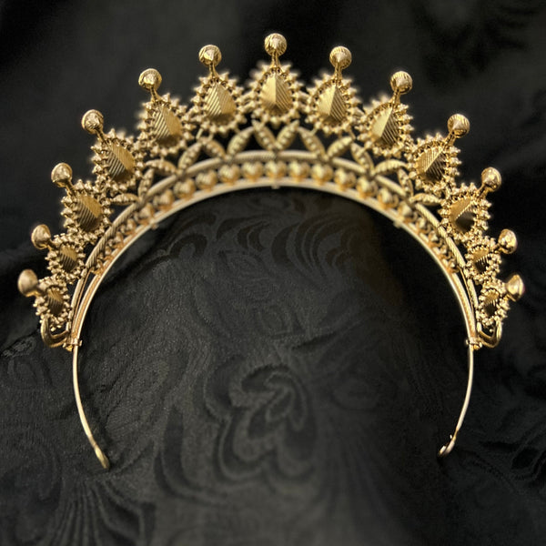 An ornate gold tiara with intricate filigree detailing and small bead accents, displayed on a textured black fabric background.