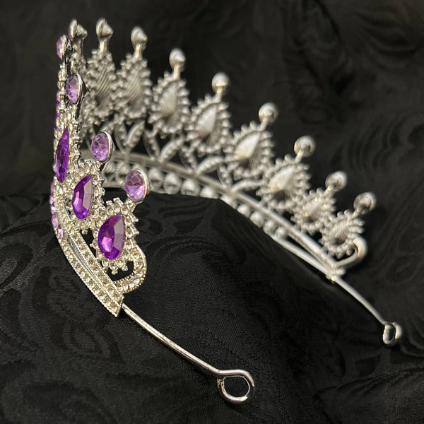 A silver tiara-style hair claw clip decorated with clear rhinestones and purple gemstones, displayed on a textured black fabric background.
