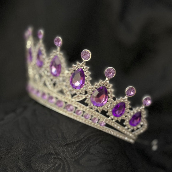 A silver tiara crown adorned with purple gemstones, resting on a textured black fabric background.