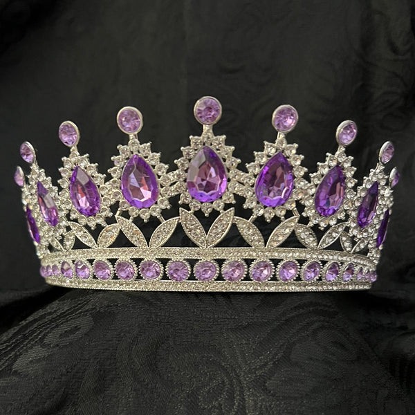 A silver tiara crown embellished with purple gemstones, displayed on a textured black fabric background.