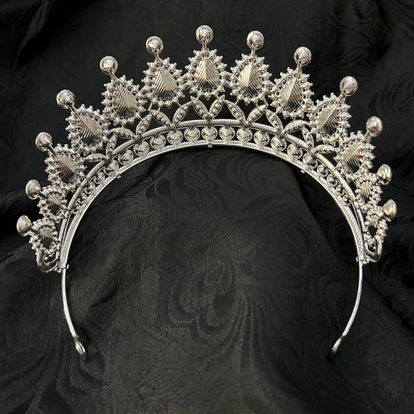 A silver filigree tiara with ornate floral and teardrop details, displayed on a textured black fabric background.